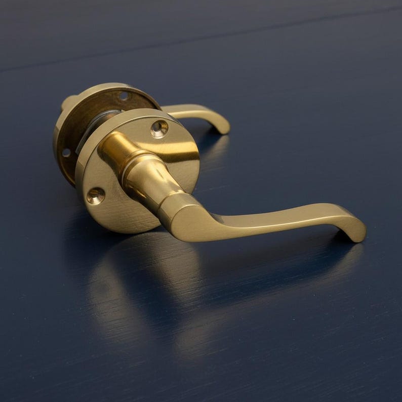 Op de afbeelding: Een messing deurklink met een gebogen hendel en een cirkelvormige achterplaat. De klink heeft een warme, gouden tint en is gemonteerd op een donkerblauwe ondergrond. Het ontwerp is eenvoudig en modern, geschikt voor binnendeuren.