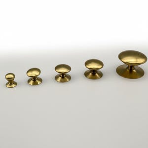 Puede incluir: Una colecci&oacute;n de cinco pomos de lat&oacute;n para armarios de diferentes tama&ntilde;os. Cada pomo presenta una parte superior redondeada y una base circular, todo con un acabado pulido. Los pomos est&aacute;n dispuestos en fila sobre un fondo blanco, mostrando sus diferentes dimensiones.