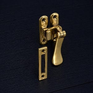 Puede incluir: Un juego de pestillo de ventana de latón, que incluye un mecanismo con bisagras y una placa de cierre separada. El pestillo tiene un mango curvo. El herraje es de color dorado brillante.