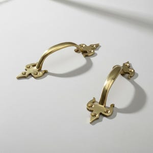 Peut inclure: Deux poignées de meuble en laiton doré avec un design incurvé. Chaque poignée a une base décorative avec un motif de style fleur de lys. Les poignées sont posées sur un fond blanc, avec des ombres ajoutant de la profondeur.