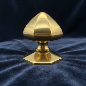 Può includere: Un pomello per armadio in ottone lucido con una parte superiore triangolare sfaccettata e una base esagonale. Il pomello ha una superficie lucida e riflettente ed è progettato per mobili o armadi. L'hardware è mostrato su uno sfondo blu scuro.