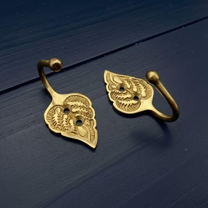 May include: Two brass leaf-shaped hooks with detailed embossed patterns. Each hook has a curved stem and a small ball at the end. The hooks are designed with a leaf pattern and are suitable for hanging items.