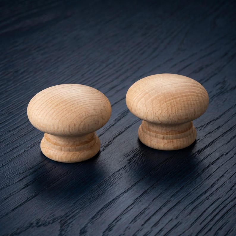 Puede incluir: Dos pomos para armario de madera natural de color claro con una parte superior lisa en forma de seta y una base circular escalonada. La veta de la madera es visible en la superficie de ambos pomos, mostrando un tono bronceado y acabado pulido.