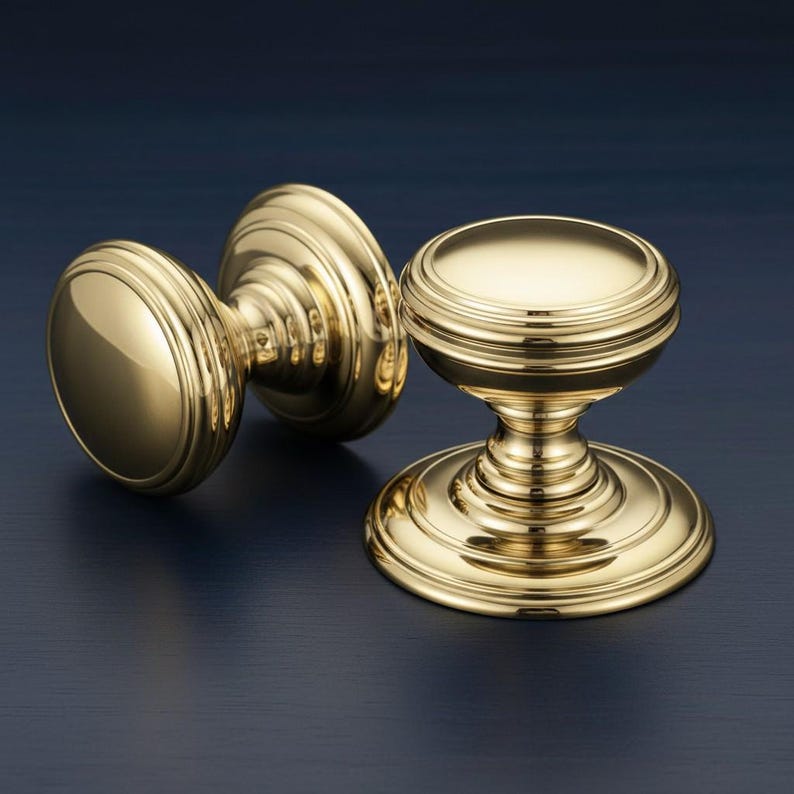 May include: Two polished brass cabinet knobs with a reflective metallic finish. Each knob features a smooth, rounded top surface and a tiered, circular base with concentric ring detailing that tapers into a central stem connecting to the knob head.