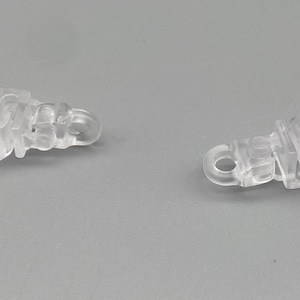 May include: Two clear plastic curtain clips, each with a hook and a hinged clip. These clips are designed for securing curtains. The clips are made of a transparent plastic material.