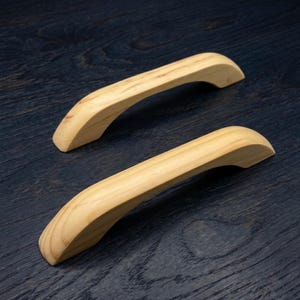 Op de afbeelding: Twee lichtgekleurde houten kastgrepen met een glad, gebogen ontwerp en zichtbare natuurlijke houtnerfpatronen. Deze hardwarestukken hebben een gebogen, ergonomische vorm met taps toelopende uiteinden en een lichtbruine afwerking.