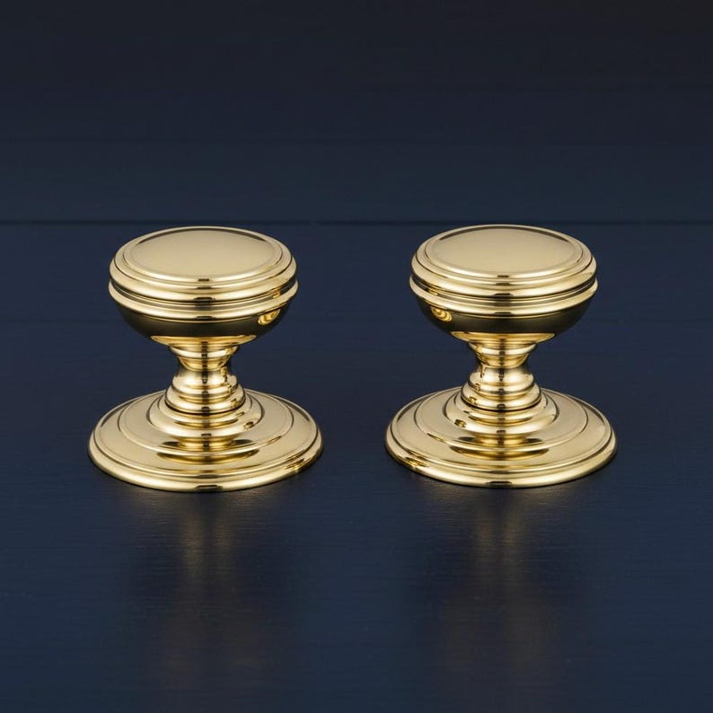 May include: Two polished brass cabinet knobs featuring a rounded top, a tiered neck, and a wide circular base. The hardware has a reflective metallic finish with concentric ring details on the top and the base, creating a uniform appearance.