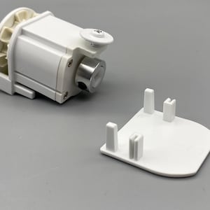 May include: A white mechanical component with a cylindrical metal piece and a separate white base with four posts. The component has a gear-like structure on one end, set against a grey background.