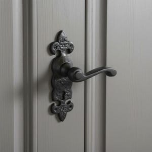 Op de afbeelding: Een matzwarte deurklinkset met een getextureerde, gehamerde metalen achterplaat met een fleur-de-lis geïnspireerde vorm aan de boven- en onderkant. De klink heeft een gebogen hendel en een sleutelgat, gemonteerd op een grijze paneeldeur.