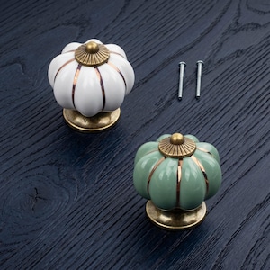 May include: Two pumpkin-shaped cabinet knobs, one white and one sage green, featuring vertical metallic copper-colored ridges and a circular antique brass-toned base and top cap. Two silver-colored metal mounting screws are included for installation.