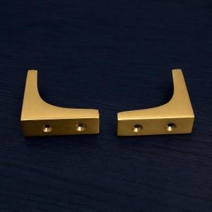 May include: Two brass-coloured, L-shaped cabinet pulls. Each pull has a rectangular base with two circular holes and a vertical extension. The pulls are displayed against a dark blue background.