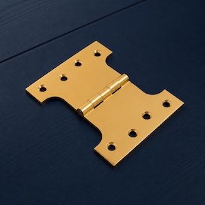 Puede incluir: Una bisagra en T dorada, un tipo de bisagra para puertas, se muestra sobre un fondo azul oscuro. La bisagra metálica tiene forma rectangular con bordes redondeados y múltiples orificios para tornillos. La bisagra está abierta, mostrando la articulación.