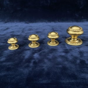 May include: Four antique brass cabinet knobs of varying sizes. Each knob features a domed top with a rope-like design and a decorative base. The knobs are arranged in a row against a dark blue background, showcasing their vintage aesthetic.
