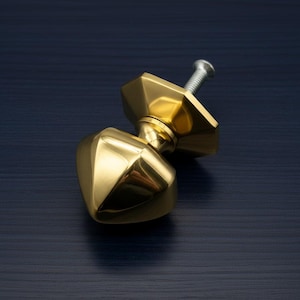 May include: A polished brass cabinet knob with a geometric design. The knob has a faceted, diamond-like shape and a threaded screw for installation. The hardware is a bright gold color.