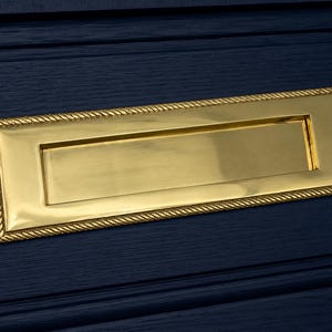 Pode incluir: Uma ranhura para cartas retangular em latão dourado com uma borda em forma de corda, montada numa porta de madeira azul escura. O latão polido reflete a luz, oferecendo um aspeto clássico e elegante.