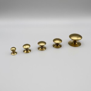 Puede incluir: Cinco pomos de lat&oacute;n para armarios de diferentes tama&ntilde;os dispuestos en fila. Cada pomo tiene una parte superior redondeada y una base circular. Los pomos son de color dorado met&aacute;lico c&aacute;lido, sobre un fondo claro.