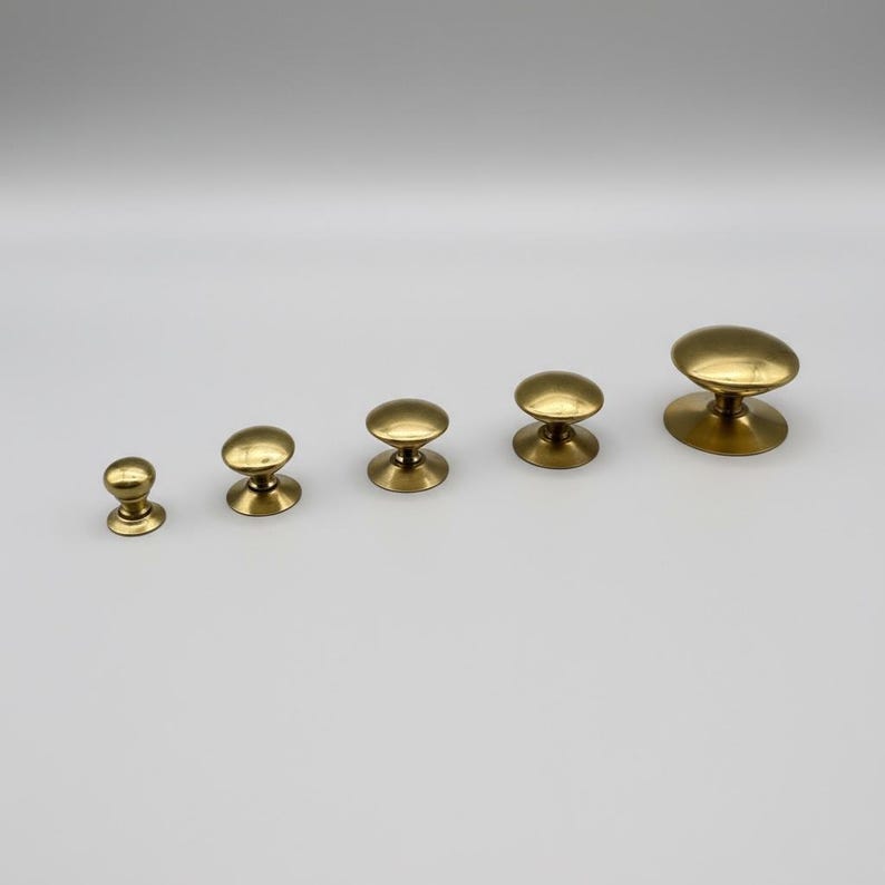Puede incluir: Cinco pomos de lat&oacute;n para armarios de diferentes tama&ntilde;os dispuestos en fila. Cada pomo tiene una parte superior redondeada y una base circular. Los pomos son de color dorado met&aacute;lico c&aacute;lido, sobre un fondo claro.