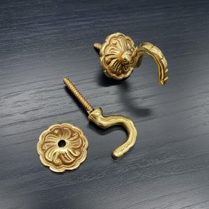 May include: Two ornate, gold-coloured metal hooks with matching decorative screw-on rosettes. One hook is attached to a screw, the other is separate. The rosettes have a floral design.