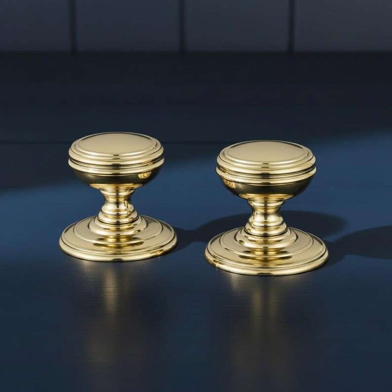 May include: Two polished brass cabinet knobs featuring a round, flat top surface with concentric circular detailing, a tapered neck, and a wide, circular base. The hardware has a reflective, metallic gold finish and a smooth, uniform texture.