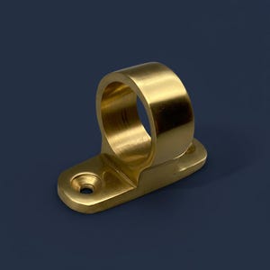 Puede incluir: Un soporte de tubería de color latón con un anillo circular y una base rectangular. El soporte tiene un solo orificio para el montaje. El metal tiene un acabado pulido, que refleja la luz.