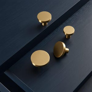 May include: Four gold-colored cabinet knobs with a brushed finish are arranged on a dark blue surface. The knobs are circular with a short, cylindrical base. The lighting highlights the metallic sheen of the hardware.