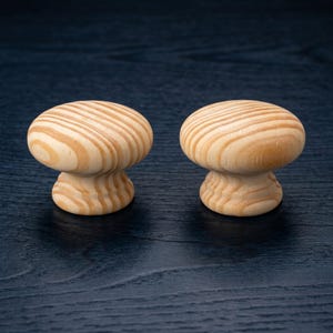 May include: Two round cabinet knobs made of light-coloured wood with a visible natural grain pattern. Each knob features a smooth, mushroom-shaped top and a tapered base, showcasing the organic texture and tan tones of the unfinished wood material.
