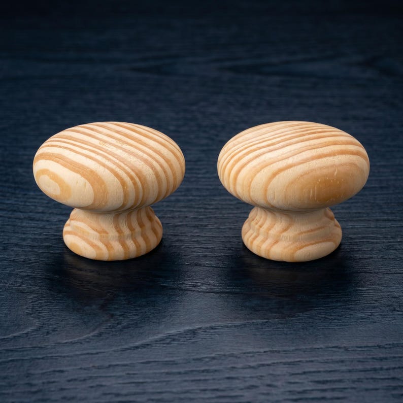 May include: Two round cabinet knobs made of light-coloured wood with a visible natural grain pattern. Each knob features a smooth, mushroom-shaped top and a tapered base, showcasing the organic texture and tan tones of the unfinished wood material.