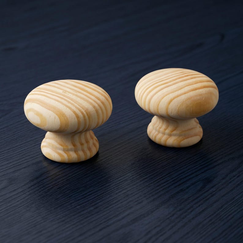 May include: Two round cabinet knobs made of light-coloured natural wood. Each knob features a smooth, mushroom-shaped top with a visible grain pattern and a tiered, circular base that tapers inward before widening at the bottom for attachment.