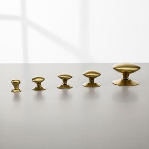 Puede incluir: Cinco pomos de lat&oacute;n de diferentes tama&ntilde;os est&aacute;n dispuestos en fila. Cada pomo tiene una parte superior redondeada y una base corta y cil&iacute;ndrica. Los pomos son de color dorado c&aacute;lido y est&aacute;n sobre un fondo neutro.