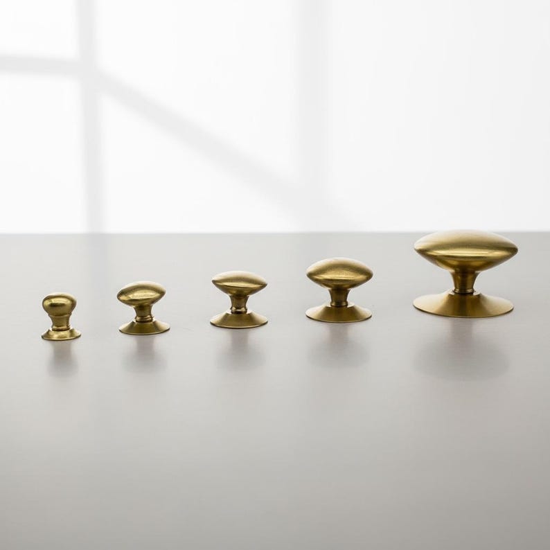 Puede incluir: Cinco pomos de lat&oacute;n de diferentes tama&ntilde;os est&aacute;n dispuestos en fila. Cada pomo tiene una parte superior redondeada y una base corta y cil&iacute;ndrica. Los pomos son de color dorado c&aacute;lido y est&aacute;n sobre un fondo neutro.