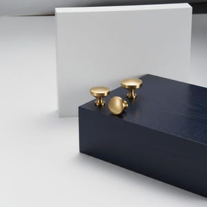 May include: Three brass cabinet knobs with a rounded design are displayed on a dark blue wooden block. The knobs have a polished finish and are arranged in a group. A white rectangular shape is in the background.