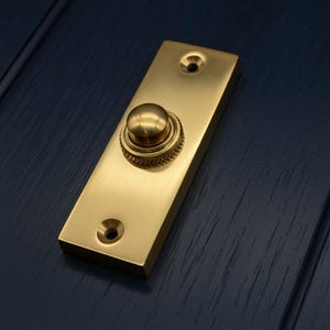 Può includere: Un pulsante per campanello rettangolare in ottone con un pulsante rotondo e due fori per le viti. Il pulsante ha un anello strutturato attorno alla base. La finitura è di colore oro lucido.