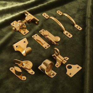 May include: An assortment of polished brass hardware components, such as handles, latches, and hinges, displayed on a dark green velvet background. The golden hardware reflects light. The collection suggests a vintage or antique home decor theme.