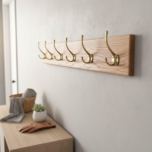 May include: A wooden coat rack featuring six brass hooks attached to a light-coloured wooden board. The hooks are evenly spaced and curved. The rack is mounted on a light grey wall. The rack's dimensions are approximately 76 cm in length.