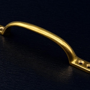 Peut inclure: Une poignée de meuble incurvée, de couleur laiton, avec une poignée arquée lisse. La poignée comporte des plaques de montage rectangulaires à chaque extrémité, chacune avec deux trous de vis. La quincaillerie est posée sur un fond bleu foncé.