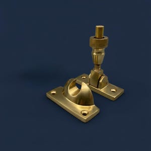May include: Two brass window stays against a dark blue backdrop. One features a curved hook, the other an adjustable arm with a threaded knob. Both have rectangular mounting plates with screw holes.