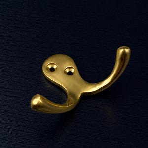 Könnte beinhalten: Ein polierter Messing-Wandhaken mit geschwungenem Design. Der Haken hat ein abgerundetes Ende und eine flache Basis mit zwei Schraubenlöchern zur Montage. Das Metall hat eine reflektierende Oberfläche, und die Form ähnelt einem stilisierten Fragezeichen.
