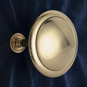 May include: A polished brass cabinet knob with a circular design. The knob has a rounded face and a decorative base, attached to a dark stem. The hardware is set against a dark blue background.