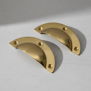 Könnte beinhalten: Zwei polierte Schubladengriffe aus Messing in Halbmondform. Jeder Griff hat ein gebogenes Design mit drei Schraubenlöchern zur Befestigung. Die Griffe sind goldfarben und reflektieren das Licht.