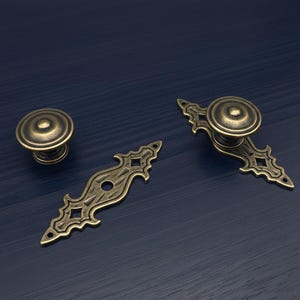 Può includere: Set di ferramenta per mobili in ottone antico. Include un pomello rotondo, una maniglia decorativa e una maniglia con pomello. La ferramenta presenta un design vintage ornato con intagli a forma di diamante e finitura in ottone scuro.