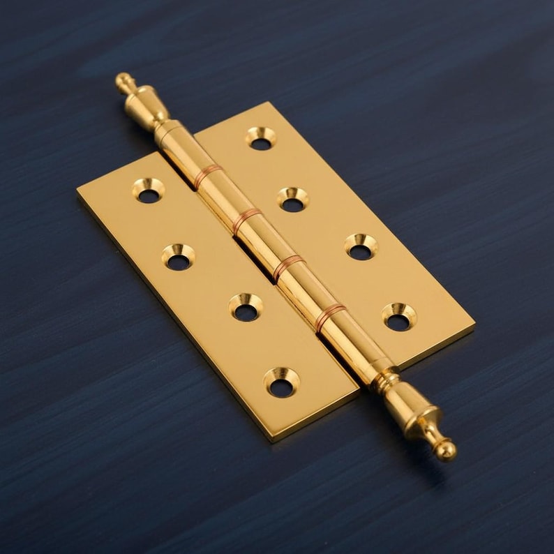 May include: A polished brass door hinge with a decorative finial. The hinge has a rectangular base with multiple screw holes and a cylindrical barrel with decorative rings. The overall design is symmetrical and ornate.