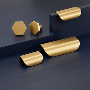 May include: A selection of gold-coloured cabinet hardware, featuring two hexagonal knobs and several curved pull handles. The hardware is presented on a dark blue surface, contrasting with the metallic finish. The pulls have a curved design, adding a modern touch.