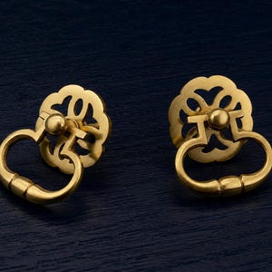 May include: Two gold-colored drawer pulls with a floral design and a curved handle. The pulls are made of metal and have a shiny finish. The background is a dark blue color.