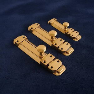 Könnte beinhalten: Drei Messing-Türriegel in verschiedenen Größen liegen auf einem dunkelblauen, strukturierten Hintergrund. Jeder Riegel hat einen rechteckigen Körper mit einem abgerundeten Knopf. Diese Riegel sind für einen sicheren Türverschluss konzipiert.