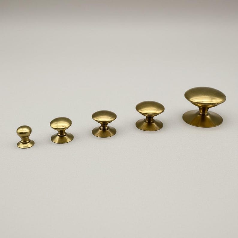Puede incluir: Cinco pomos de lat&oacute;n para armarios de diferentes tama&ntilde;os dispuestos en fila. Cada pomo tiene una parte superior redondeada y una base acampanada, con un acabado met&aacute;lico c&aacute;lido. Los pomos est&aacute;n sobre un fondo liso y claro.