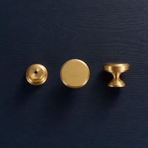 May include: Three brass cabinet knobs in various designs are displayed against a dark blue wooden background. The knobs include a flat disc, a round knob, and a decorative knob with a flared base.