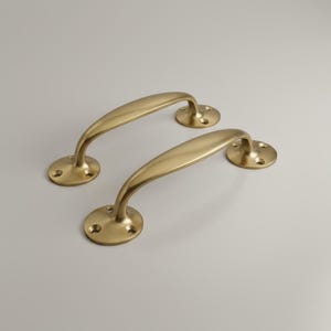 Peut inclure: Deux poignées de porte de couleur laiton. Chaque poignée a une poignée incurvée et une base circulaire avec des trous de vis. Le matériel a une finition lisse et polie et un design classique. Les poignées sont probablement pour une armoire ou une porte.