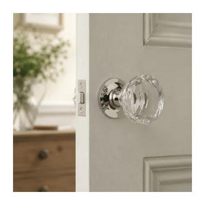 Peut inclure: Un bouton de porte en verre transparent à facettes avec une plaque de base circulaire en chrome poli et une gâche assortie, monté sur une porte blanche à panneaux. Le bouton présente une taille en diamant à facettes multiples qui reflète la lumière.