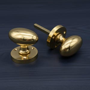 Peut inclure: Deux poignées de porte en laiton poli de forme ovale. Chaque bouton a une base ronde avec des trous de vis. L'un des boutons est fixé à une tige et à une plaque circulaire. Les boutons sont posés sur un fond bleu foncé.