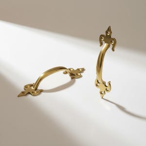 Puede incluir: Dos tiradores dorados para armarios con un diseño curvo y extremos decorativos. Los tiradores son de metal y están sobre un fondo blanco con sombras. Los tiradores miden aproximadamente 15 cm de largo.